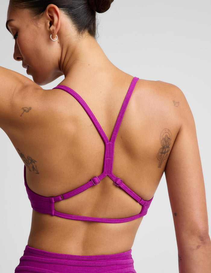 A woman shown from the back wearing a bright magenta-purple sports bra with crisscrossing underarm straps and a matching high-waisted athletic bottom against a light gray studio background.