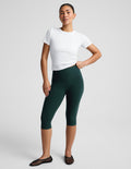 A woman modeling dark green high-waisted capri leggings with a white t-shirt and black flat shoes, standing against a light gray studio background.