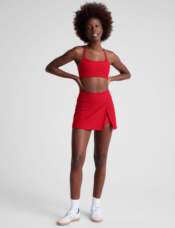 A woman wears the red Spacedye Points Crossover Skirt with a matching sports bra, featuring an asymmetrical wrap front slit.