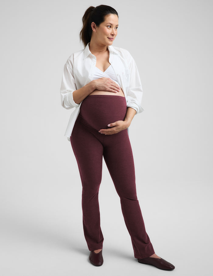 A pregnant woman wearing a white nursing bra, white button-up shirt, and red bootcut maternity leggings against a plain studio background.