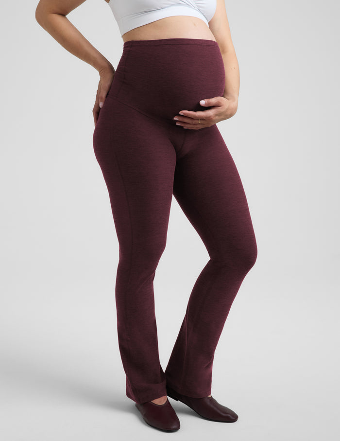 A pregnant woman wearing a white nursing bra and red bootcut maternity leggings against a plain studio background.