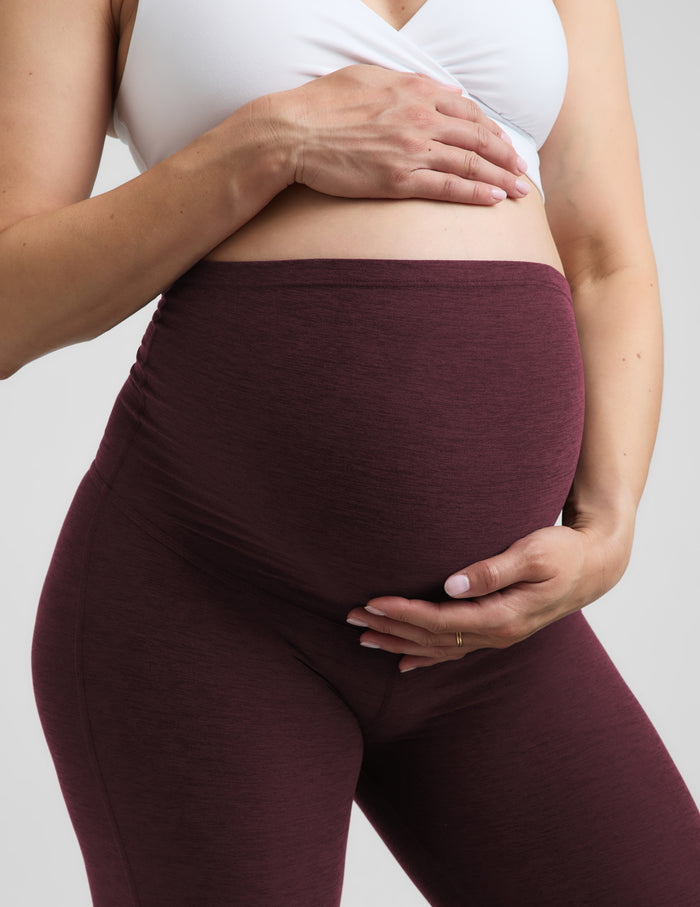 A pregnant woman wearing a white nursing bra and red bootcut maternity leggings against a plain studio background.