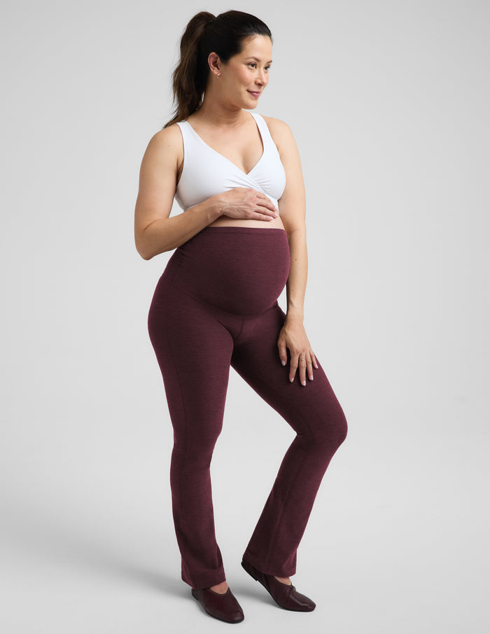 A pregnant woman wearing a white nursing bra and red bootcut maternity leggings against a plain studio background.