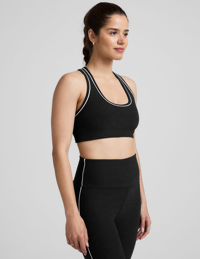 model wearing a black scoop neck racerback sports bra and leggings with white trim on a gray background.