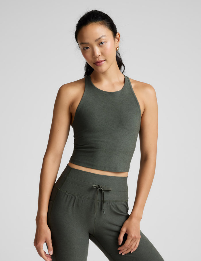 Woman is wearing a green racerback high-neck cropped tank and joggers against a plain background.