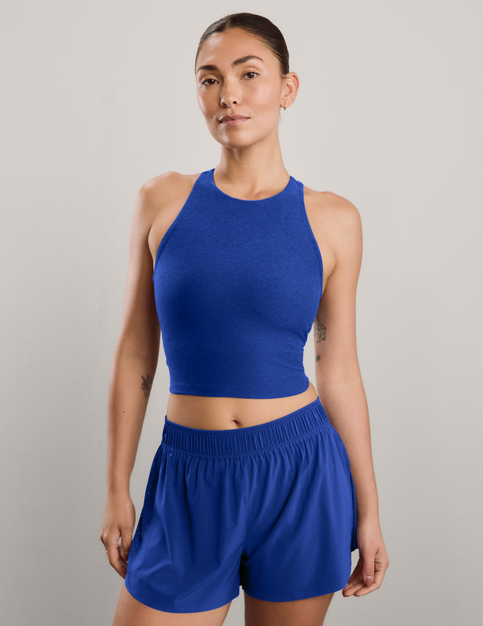 A woman modeling a matching athletic set consisting of a royal blue high neck crop top and matching high-waisted shorts against a neutral gray studio background.