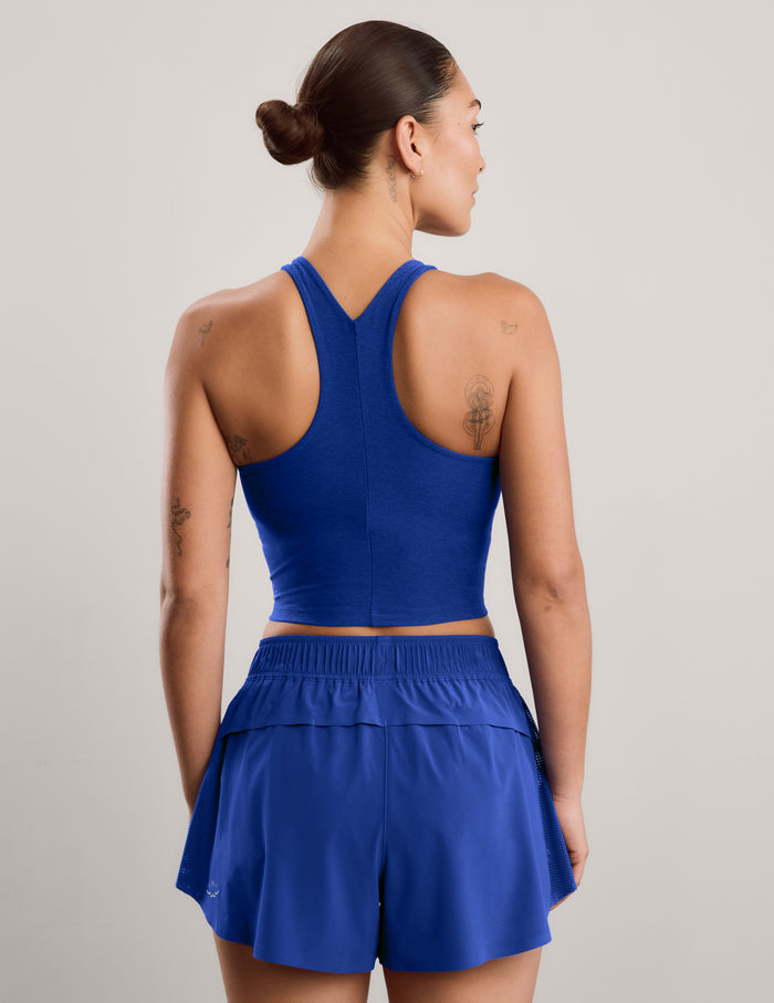 A woman modeling a matching athletic set consisting of a royal blue high neck crop top and matching high-waisted shorts against a neutral gray studio background.