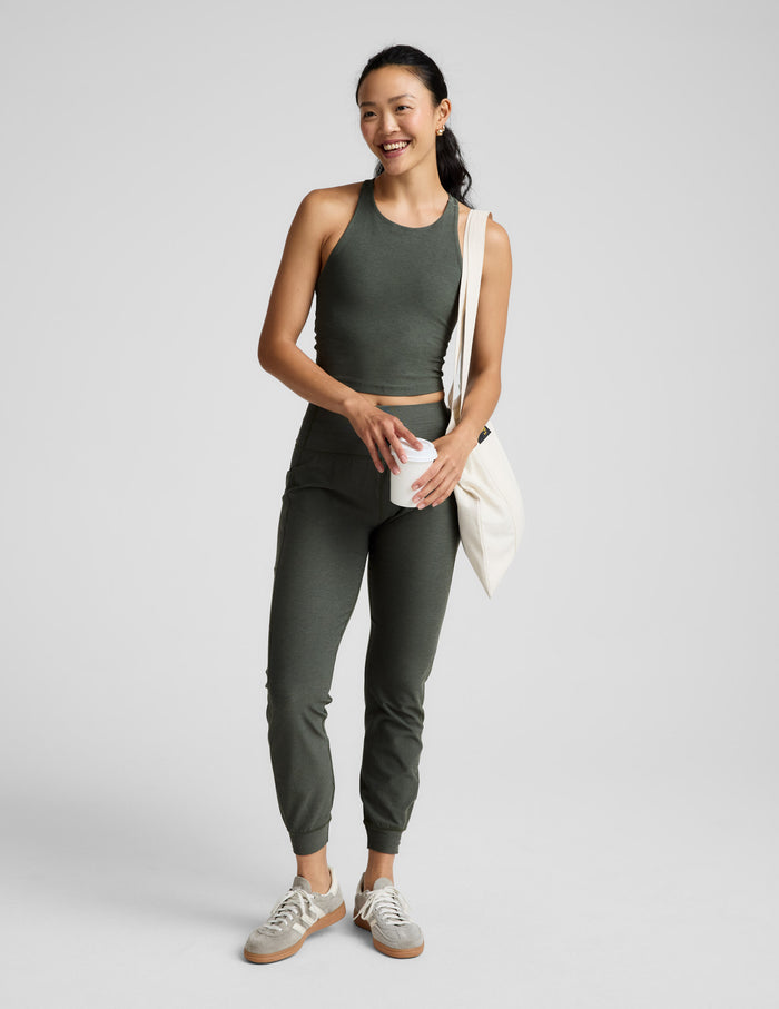 Woman is wearing a green racerback high-neck cropped tank and joggers against a plain background.