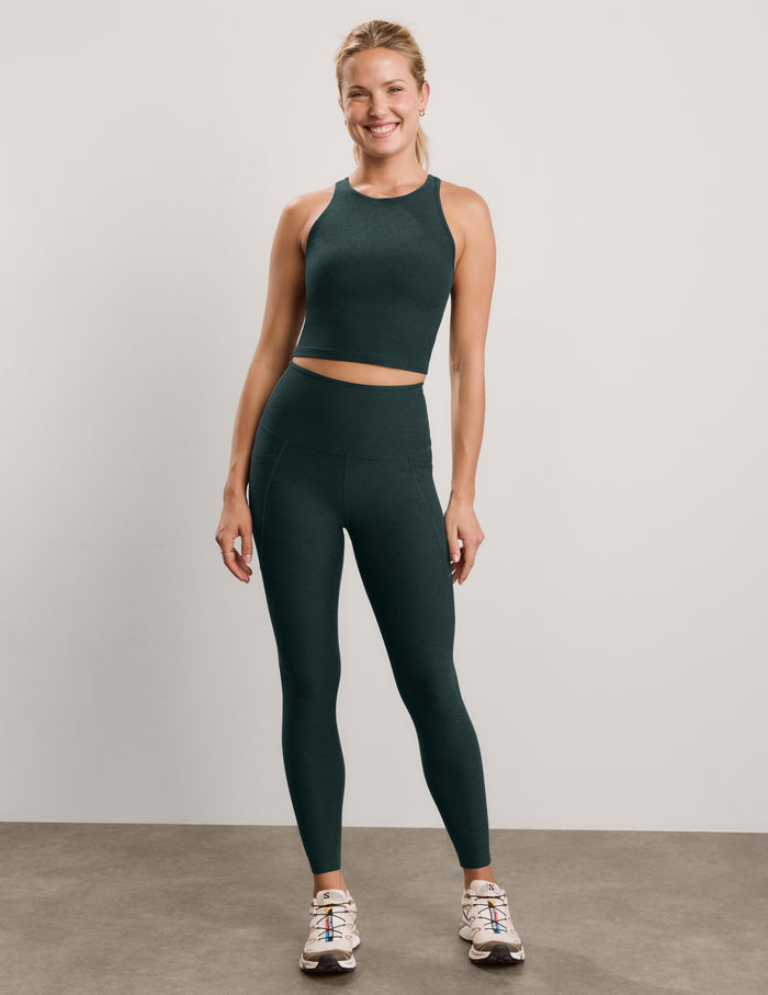 A smiling blonde woman posing in a dark forest green athletic crop top and matching high-waisted leggings against a light gray studio background.