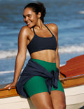 A woman wearing a dark navy sports bra and green athletic shorts, sitting on the edge of a wooden boat with the ocean in the background.