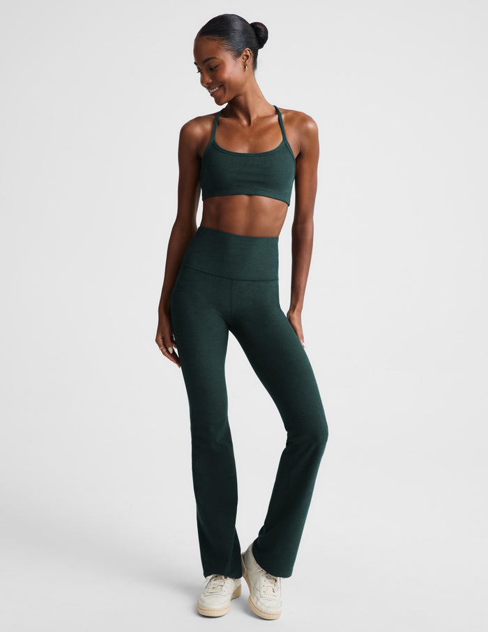 A woman modeling a matching dark green activewear set consisting of a sports bra and high-waisted bootcut leggings, paired with white sneakers, against a light gray studio background.