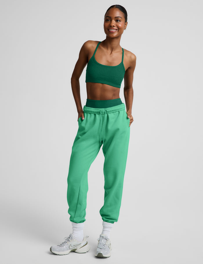 Woman wearing a green sports bra and green biker shorts with light green sweatpants on top against a plain background.