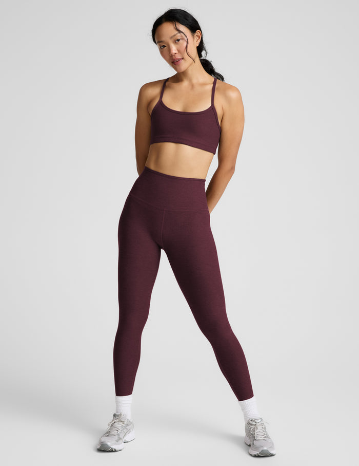 A woman modeling a matching athletic set consisting of a scoop-neck sports bra and high-waisted leggings in a deep red color, shot against a solid light gray background.
