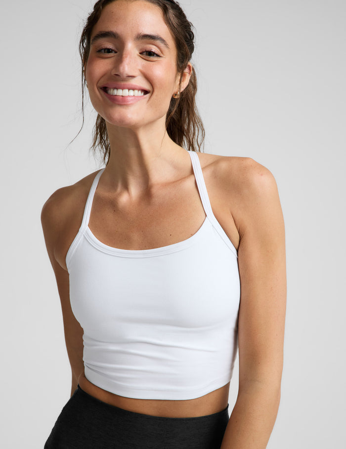 A smiling woman wearing a white crop top tank with thin shoulder straps, paired with black bottoms, photographed against a light gray studio background.