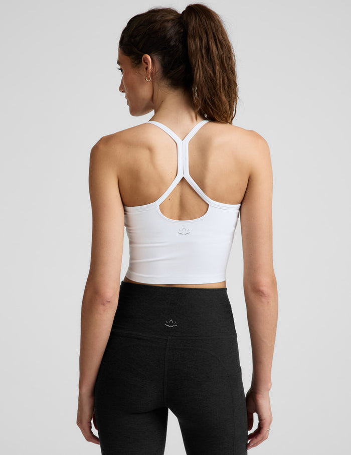 A smiling woman wearing a white crop top tank with thin shoulder straps, paired with black bottoms, photographed against a light gray studio background.