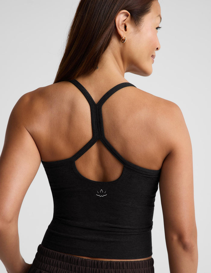 A woman shown from the back wearing a black knit camisole tank top and dark brown pants, standing against a light gray studio background.
