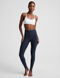 A woman modeling high-waisted navy blue leggings and a white sports bra against a light gray studio background.