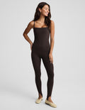 A woman modeling a dark brown, sleeveless, full-length stirrup bodysuit or jumpsuit against a light gray studio background.