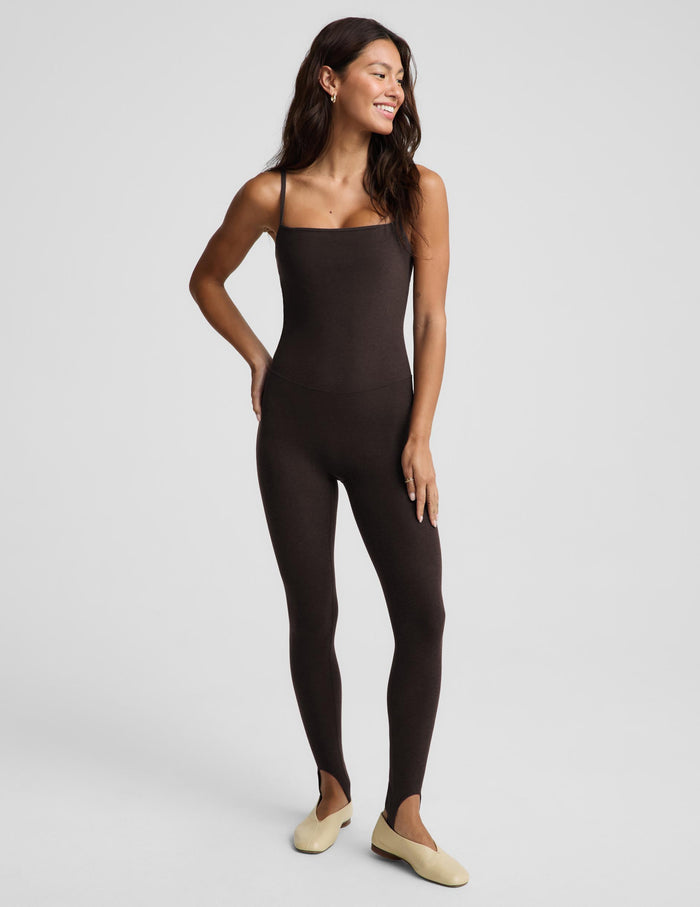 A woman modeling a dark brown, sleeveless, full-length stirrup bodysuit or jumpsuit against a light gray studio background.