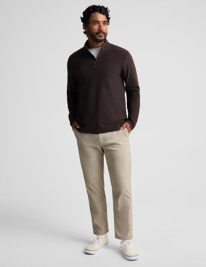 A man wearing a dark brown quarter-zip long-sleeve knit pullover over a white t-shirt and khaki pants, standing against a light gray studio background.