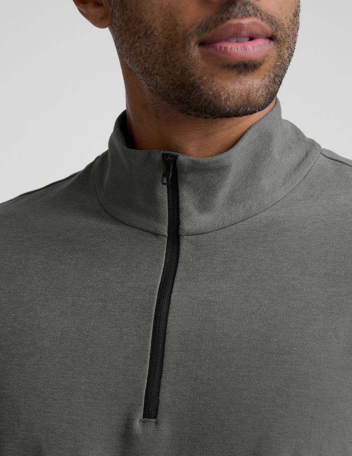 man wearing a gray quarter zip pullover with black pants and a plain background.