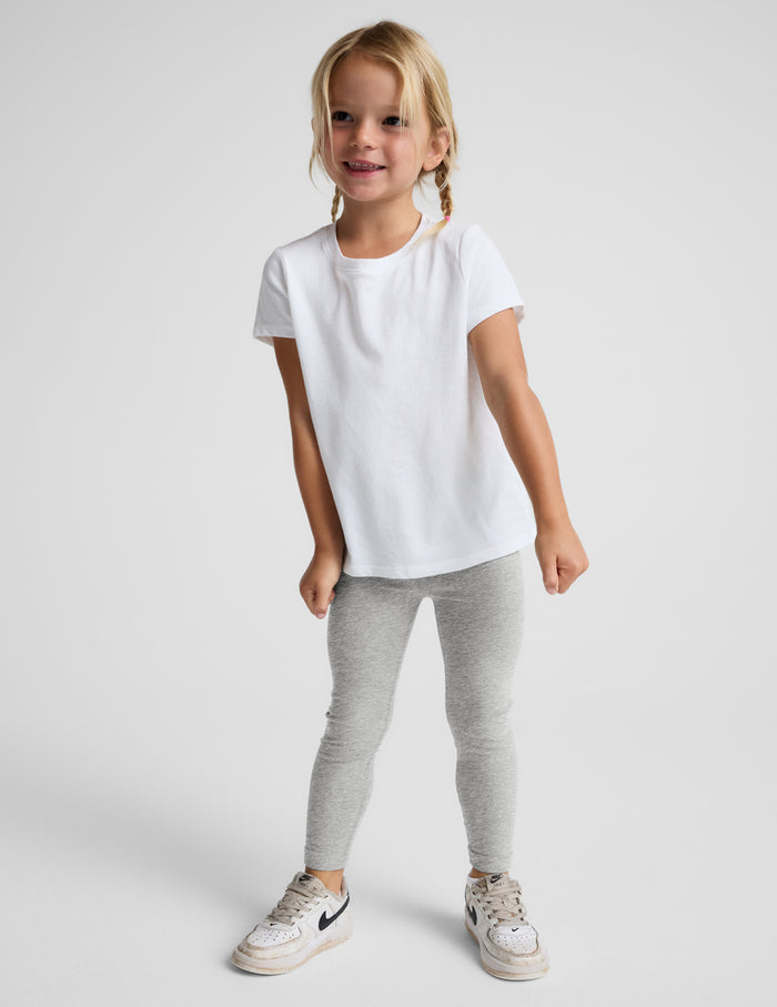 A young girl with blonde braids stands against a light gray studio background wearing a white short-sleeve t-shirt, light gray heathered leggings, and white sneakers.