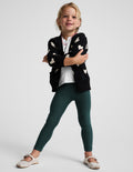 A young girl modeling a black knit cardigan with cream hearts, dark green leggings, a white t-shirt, and white Mary Jane shoes, posed against a light gray studio background.