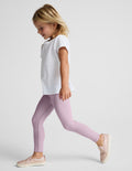 A young girl walking in profile wearing a white short-sleeve top, light purple leggings, and pale pink slip-on shoes against a light gray studio background.