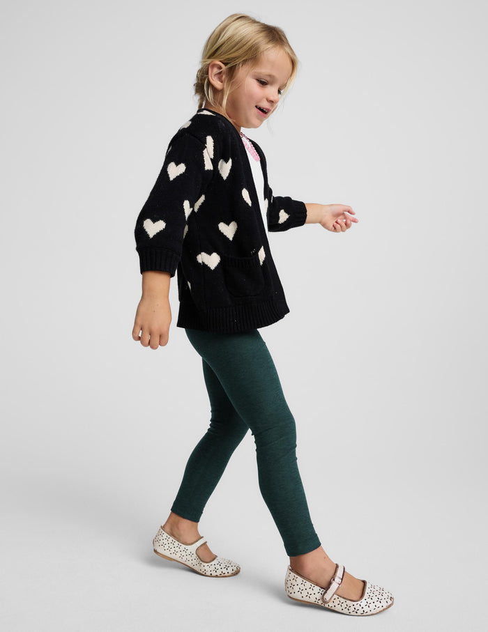 A young girl walking in profile wearing a black knit cardigan with white heart patterns, dark green leggings, and white Mary Jane shoes with a black pattern, against a light gray studio background.