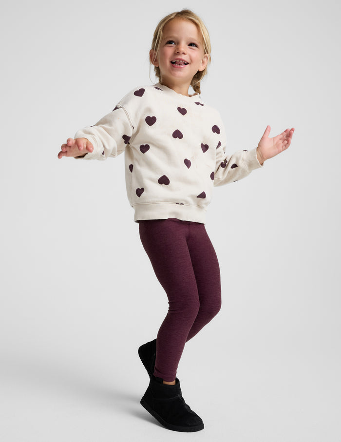 A young girl modeling a cream-colored fleece hoodie with a dark burgundy heart pattern, paired with burgundy leggings and black boots, against a light gray studio background.