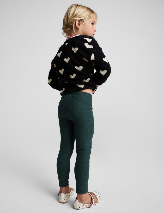 A young girl with blonde hair is shown from the back wearing a black knit sweater with a cream heart pattern and dark green leggings, standing against a light gray studio background.