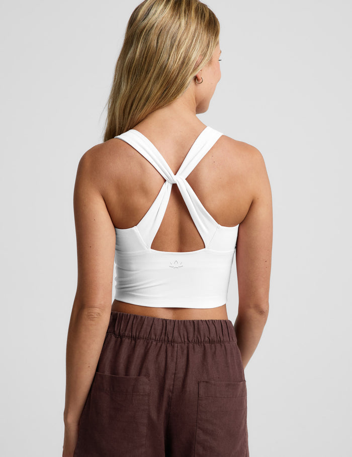 A woman shown from the back wearing a white sports bra with a crossover crisscross back strap design, paired with brown linen shorts, against a light gray studio background.