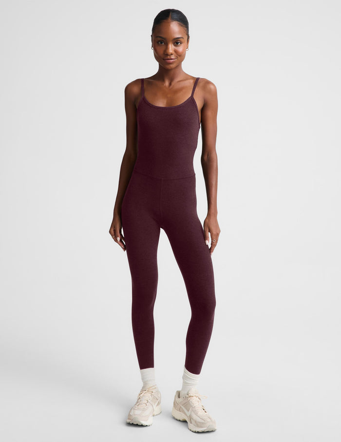 A model wearing a burgundy midi length tank jumpsuit, styled with white socks and beige sneakers, against a light gray studio background.