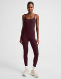 A model wearing a burgundy midi length tank jumpsuit, styled with white socks and beige sneakers, against a light gray studio background.