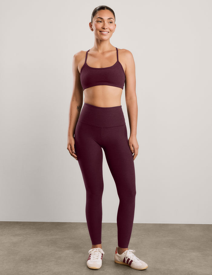 A woman modeling a matching burgundy activewear set consisting of a sports bra and high-waisted leggings, paired with white sneakers, against a neutral studio background.