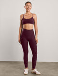 A woman modeling a matching burgundy activewear set consisting of a sports bra and high-waisted leggings, paired with white sneakers, against a neutral studio background.