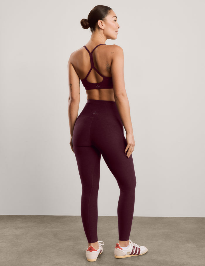 A woman modeling a matching burgundy activewear set consisting of a sports bra and high-waisted leggings, paired with white sneakers, against a neutral studio background.