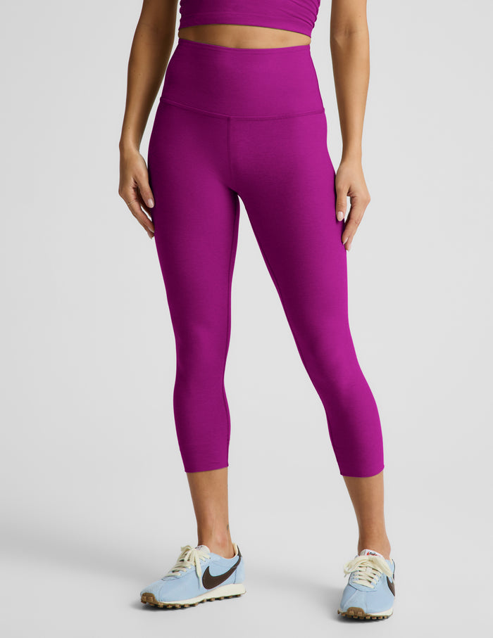 Woman wearing a purple cropped tank top and purple capri leggings against a plain background.