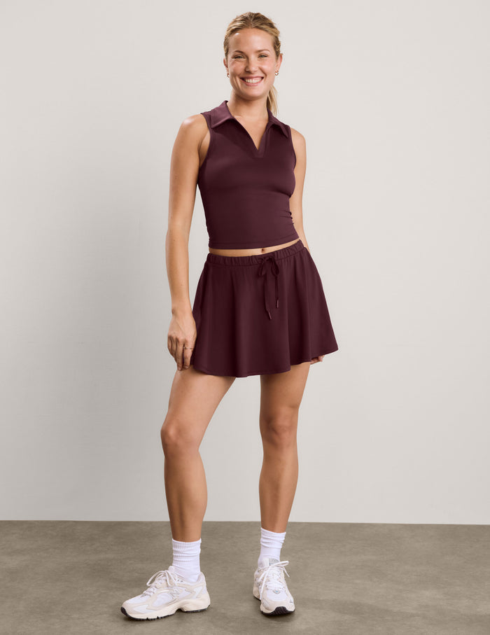 A woman modeling a sleeveless collared athletic top and matching flared mini skirt in a deep plum-burgundy color, paired with white socks and sneakers, against a neutral studio backdrop.