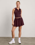 A woman modeling a sleeveless collared athletic top and matching flared mini skirt in a deep plum-burgundy color, paired with white socks and sneakers, against a neutral studio backdrop.