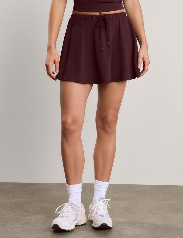 A model wearing a dark plum-colored athletic mini skirt with a drawstring waist, paired with white socks and white sneakers, standing against a neutral studio background.