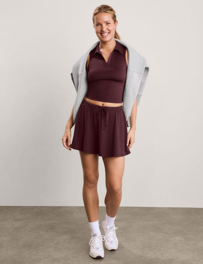 A woman modeling a dark burgundy athletic set consisting of a sleeveless collared crop top and a matching mini skirt, styled with a light gray sweatshirt draped over her shoulders, white socks, and white sneakers against a neutral studio backdrop.
