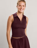 A smiling blonde woman wearing a sleeveless collared knit top in deep burgundy, paired with matching drawstring skirt, against a plain light gray studio background.