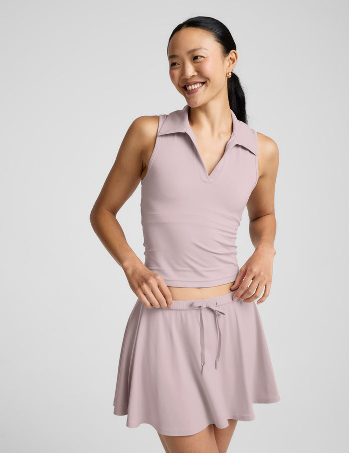 Woman wearing a light pink polo tank with a cutout in the back and a skirt against a plain background.