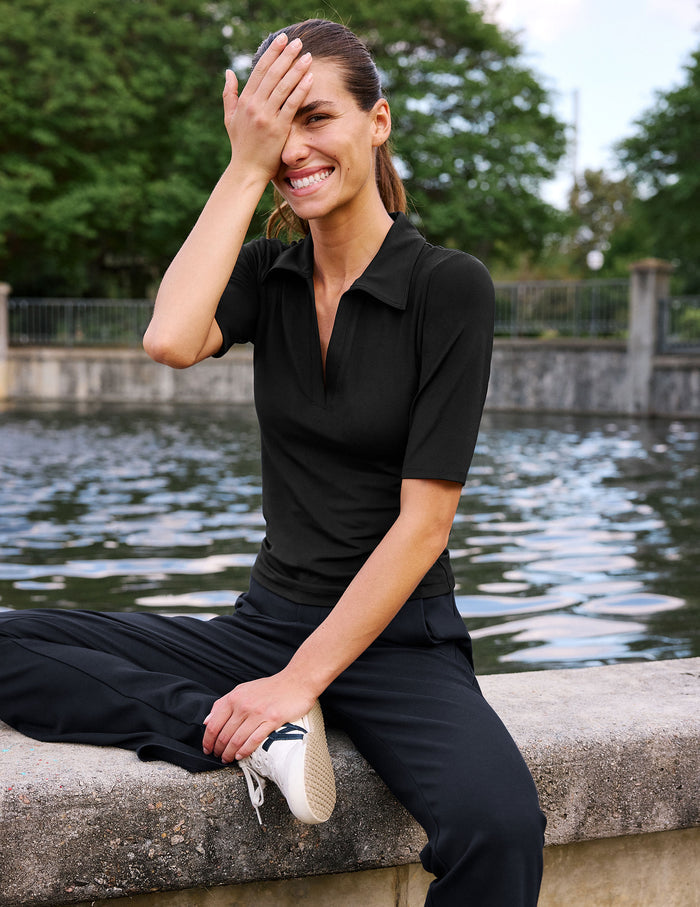 A woman sitting on a stone ledge by a body of water, wearing a fitted black short-sleeve polo shirt and black pants, with one hand covering part of her face.