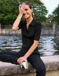 A woman sitting on a stone ledge by a body of water, wearing a fitted black short-sleeve polo shirt and black pants, with one hand covering part of her face.