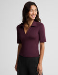 A woman wearing a short-sleeve polo shirt in deep burgundy with a split V-neckline, paired with black pants, photographed against a solid light gray studio background.