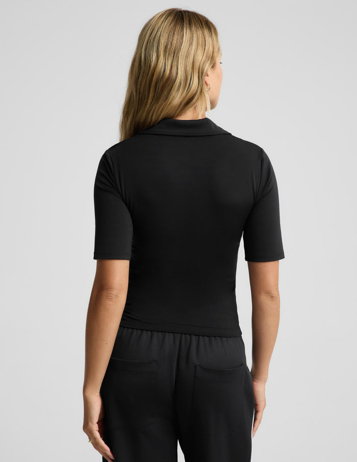 A woman modeling a black short-sleeve polo shirt with a V-neck and collar, paired with black pants, against a light gray studio background.