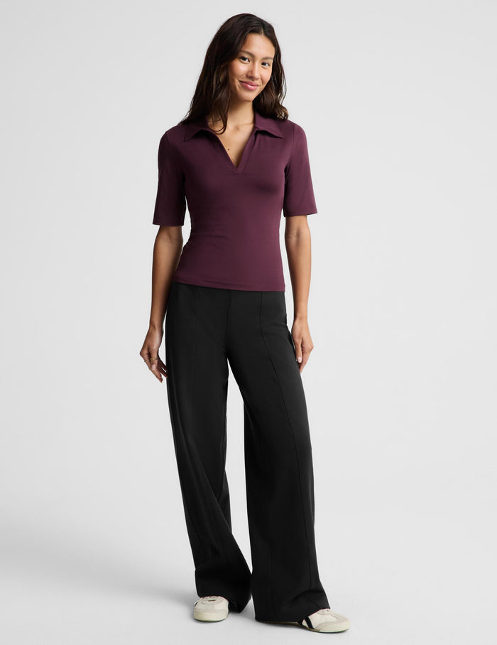 A woman wearing a short-sleeve polo shirt in deep burgundy with a split V-neckline, paired with black pants, photographed against a solid light gray studio background.