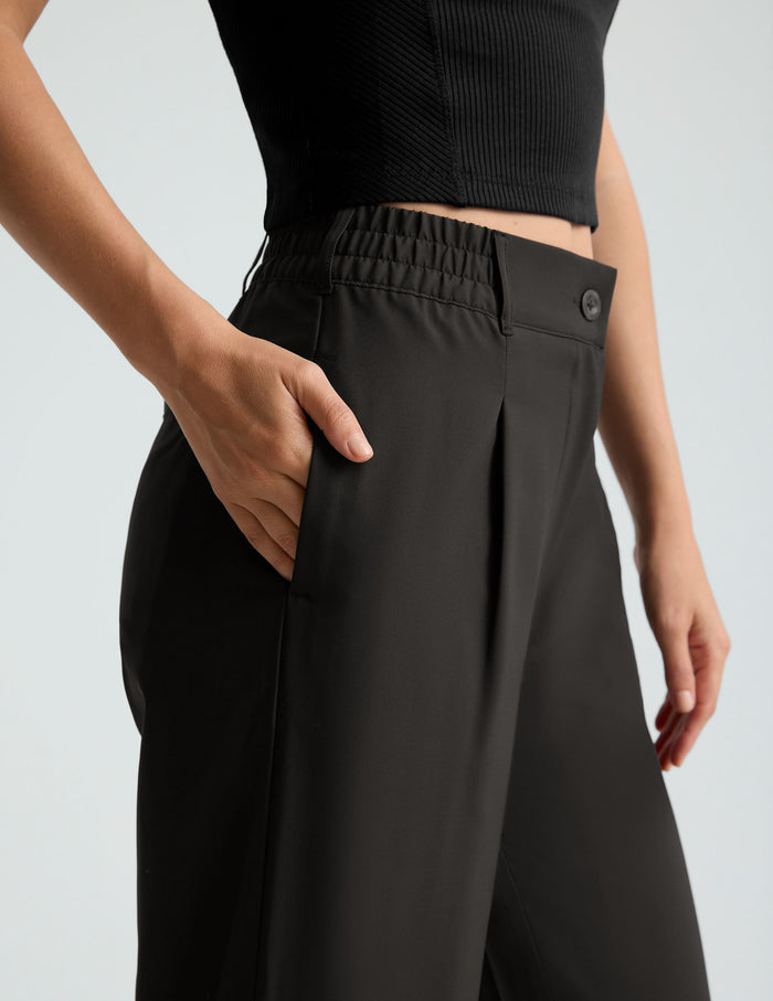 Close-up of the black Status Wide Leg Trouser showing side pockets, pleated front, and elastic waistband on a model.